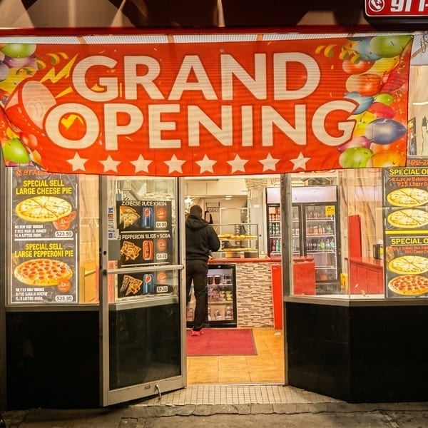 A pizza restaurant's storefront at night, with a "Grand Opening" banner and pizza specials displayed.
