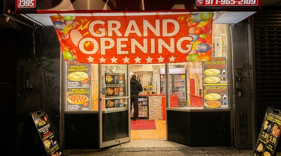 A pizza restaurant's storefront at night, with a "Grand Opening" banner and pizza specials displayed.