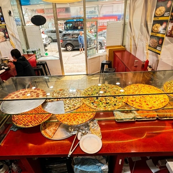 A pizza shop interior with a display case full of various pizzas and two customers at the counter.