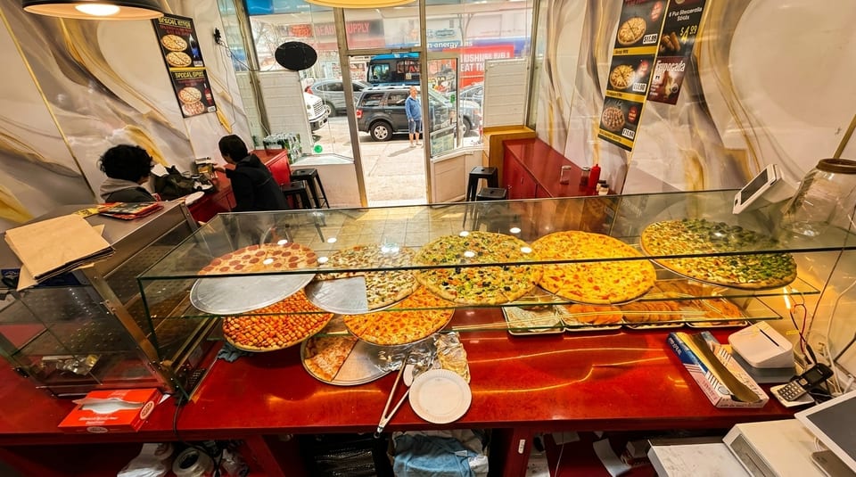 A pizza shop interior with a display case full of various pizzas and two customers at the counter.